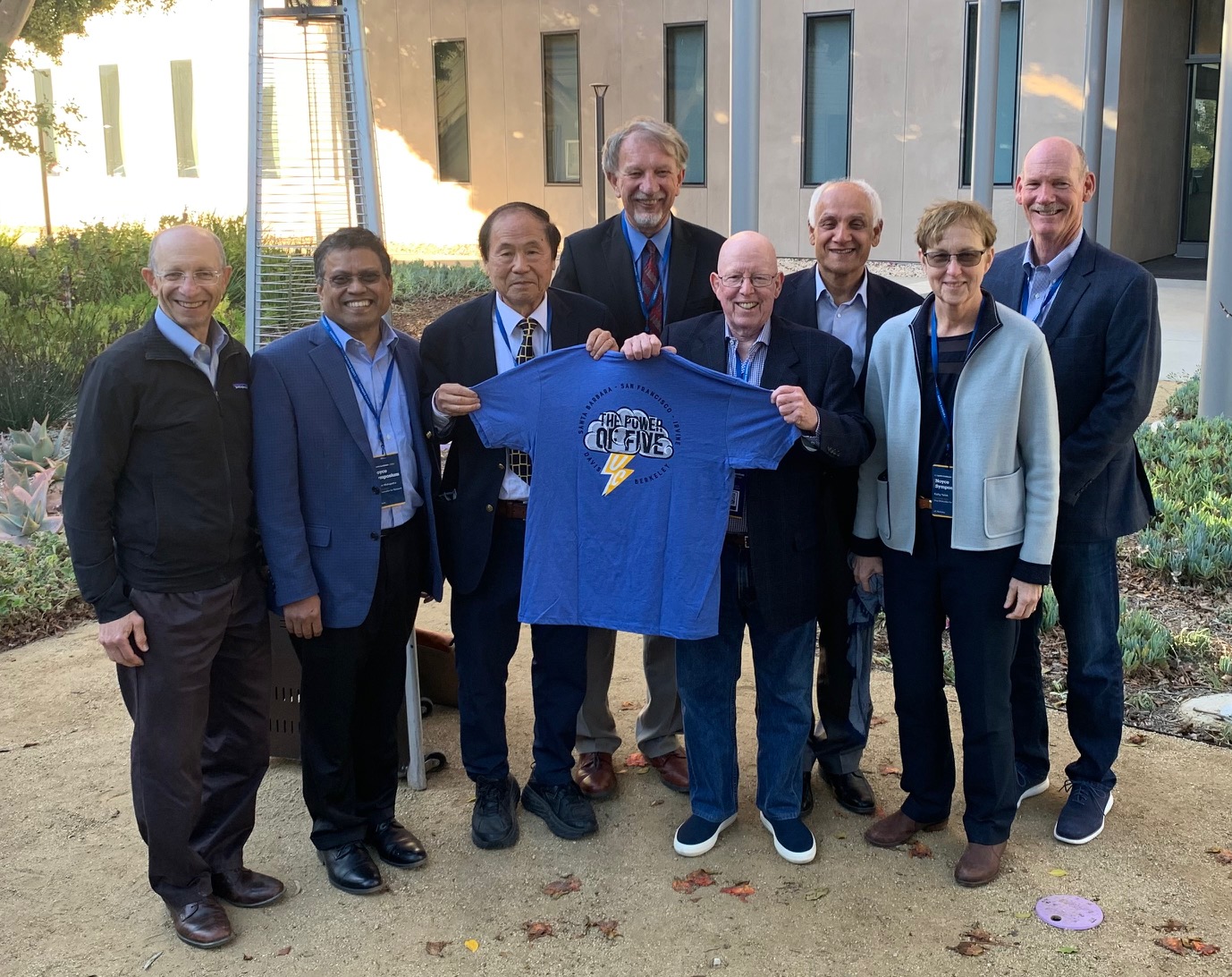 Seven gentlemen and one lady stand in a group. Asian and bald man stand in the middle holding up a t-shirt that says "The Power of Five" with a cloud and lightening bolt on it.