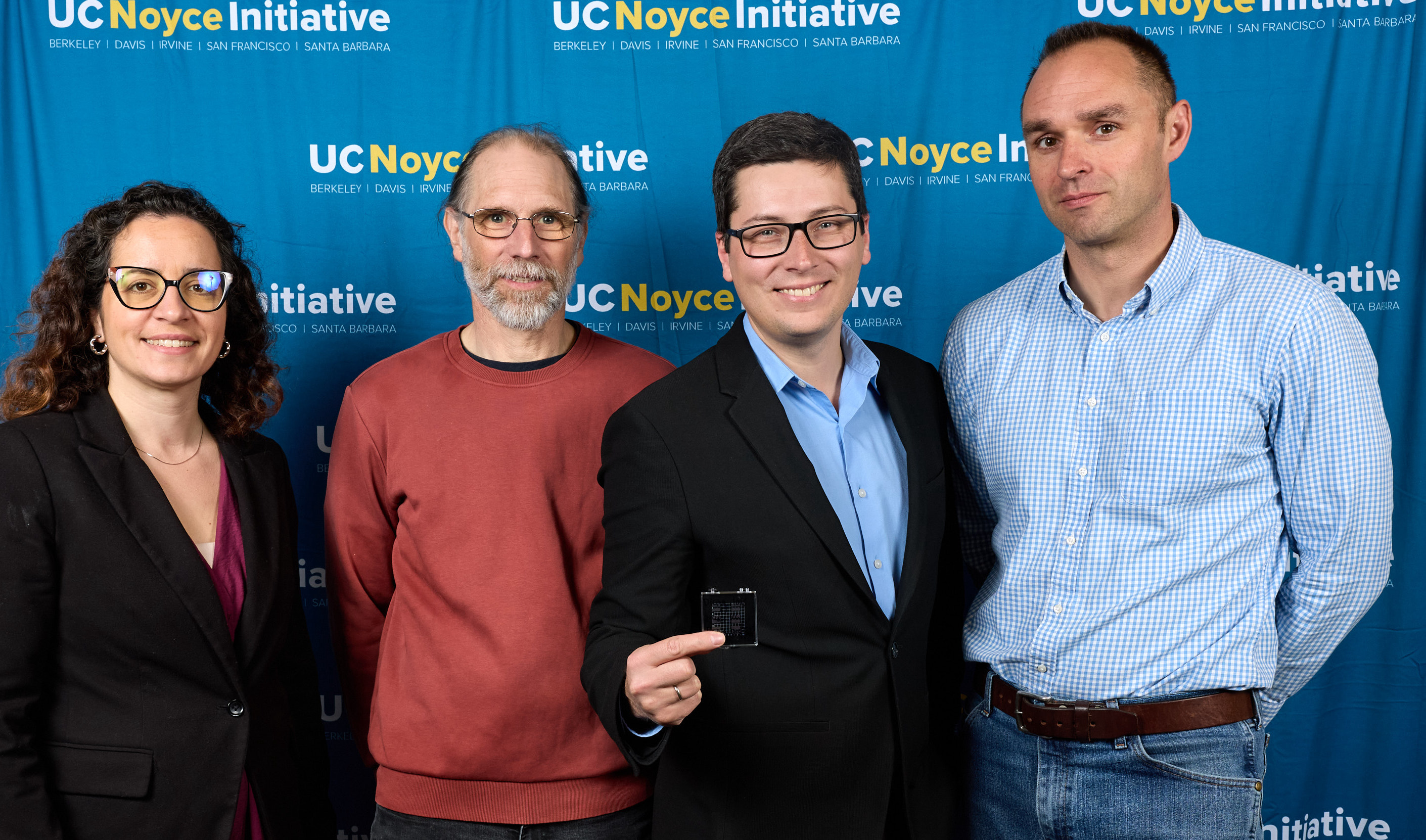 one female professor and four male professors stand together in front of UC Noyce Initiative back drop. Man in middle wearing glasses hold up quantum computer chip toward the camera.