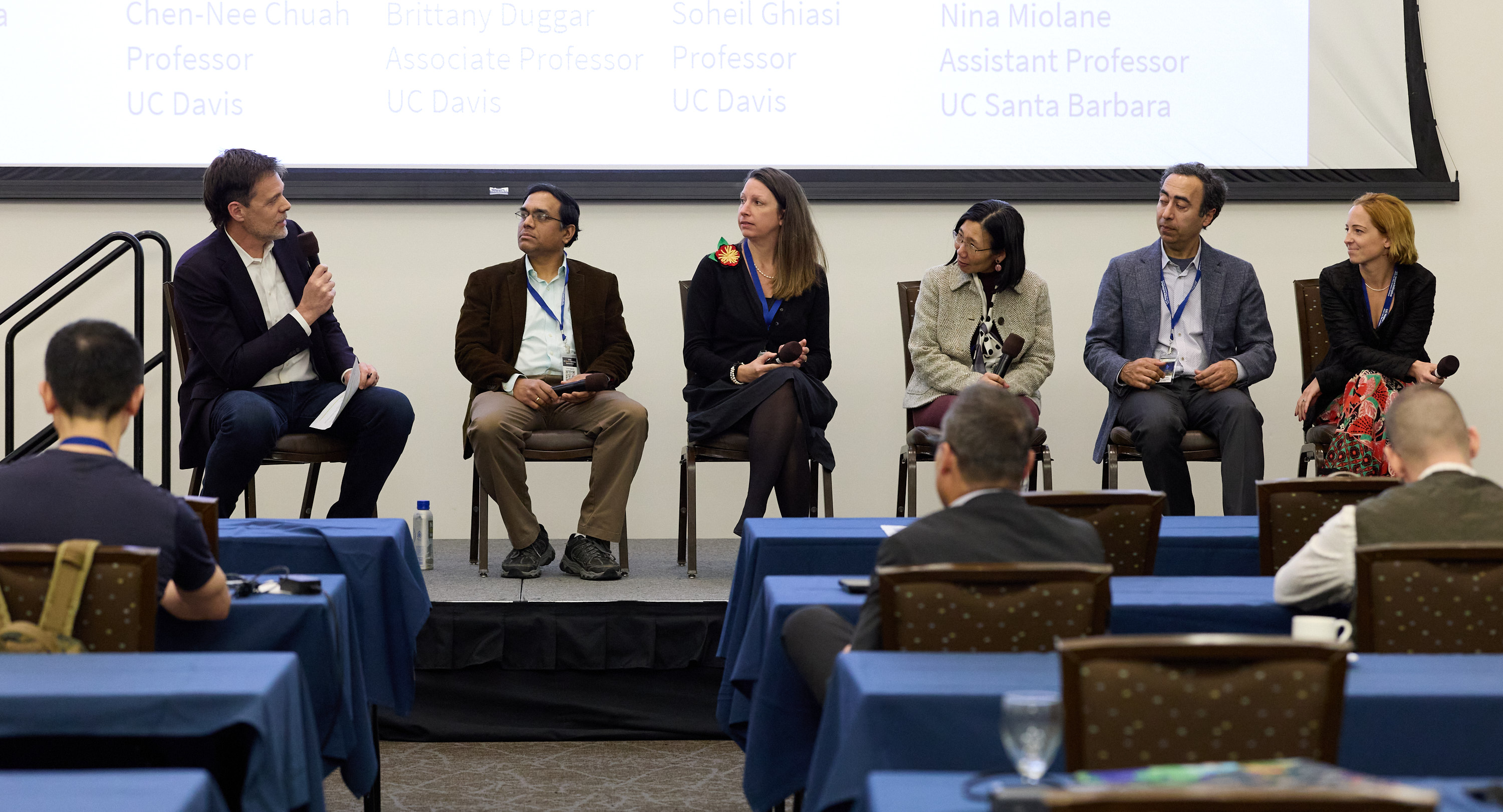 Six people sit on stage in chairs engaged in conversation in front of conference room of people. One person is speaking, five are listening.