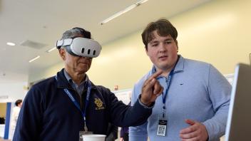 Man wearing virtual reality head set points finger in the air with a male graduate student standing nearby looking at a laptop to guide the interaction.