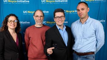 one female professor and four male professors stand together in front of UC Noyce Initiative back drop. Man in middle wearing glasses hold up quantum computer chip toward the camera.