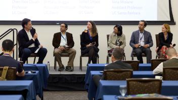 Six people sit on stage in chairs engaged in conversation in front of conference room of people. One person is speaking, five are listening.