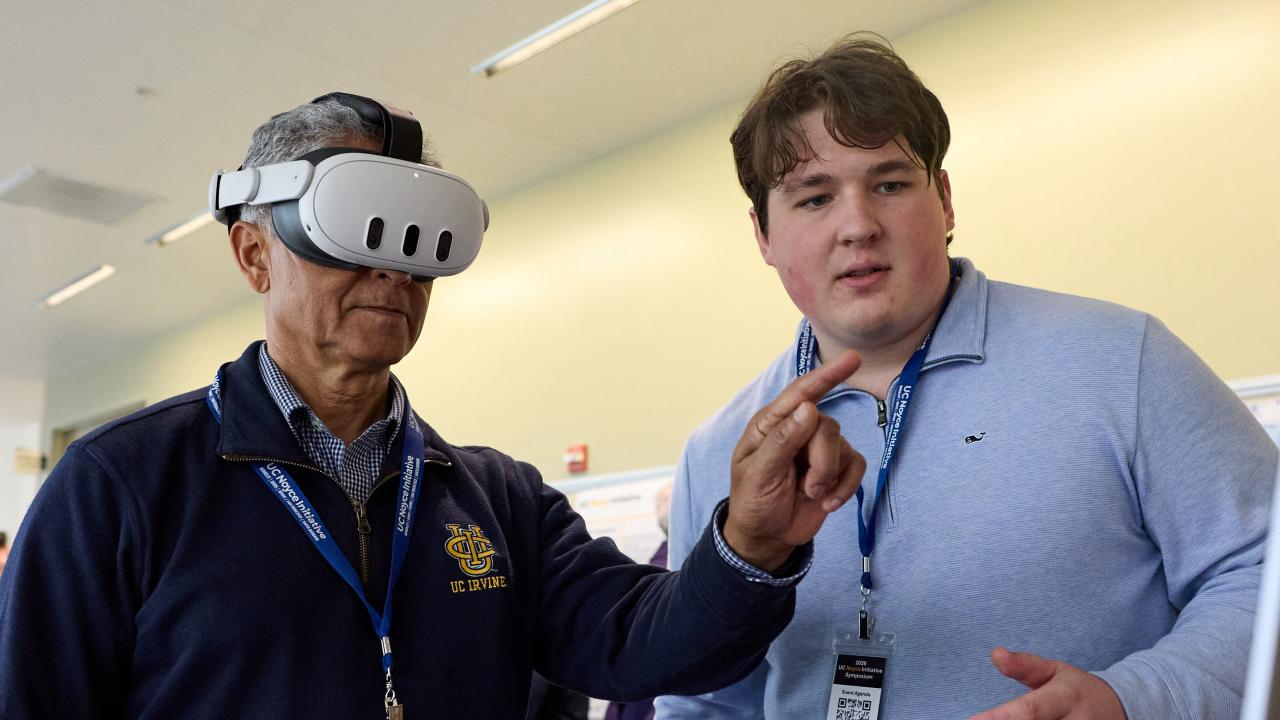 Man wearing virtual reality head set points finger in the air with a male graduate student standing nearby looking at a laptop to guide the interaction.