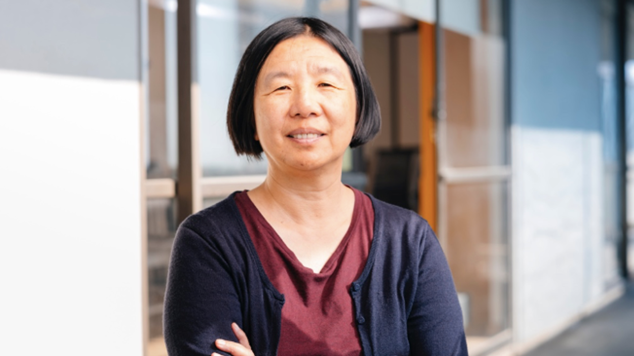 Asian woman with short hair, wearing a maroon top and a black cardigan sweater stands with her arms crossed in a well lit hallway of windows. She has a slight smile on her face.