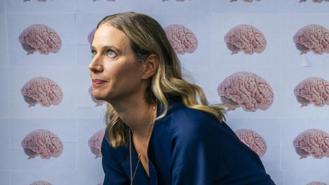 Emily Jacobs sits on a box with hands clasped resting on her knees. She is looking up off to the side of the image. Behind her and covering the box is wall paper that has rows and columns of human brains on a light blue background.