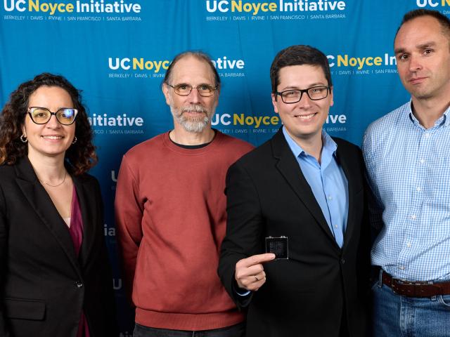 one female professor and four male professors stand together in front of UC Noyce Initiative back drop. Man in middle wearing glasses hold up quantum computer chip toward the camera.