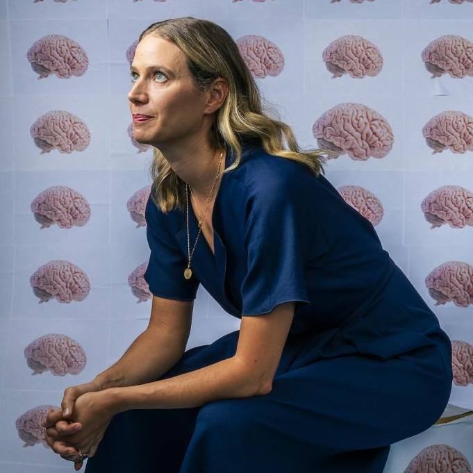 Emily Jacobs sits on a box with hands clasped resting on her knees. She is looking up off to the side of the image. Behind her and covering the box is wall paper that has rows and columns of human brains on a light blue background.