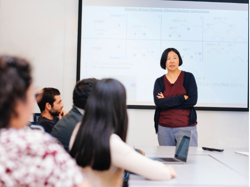 Asian woman with short hair stands with arms crossed in front of a white board talking to a class of students.