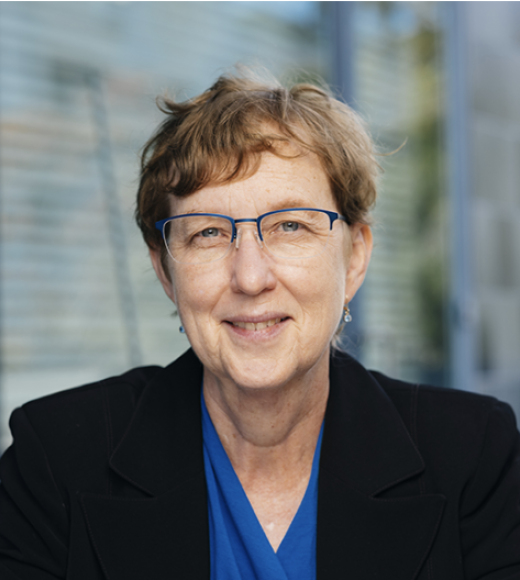 White woman with short, reddish-brown hair and blue horn-rimmed glasses smiles at camera. She is wearing a blue sports coat and a royal blue v-neck blouse.