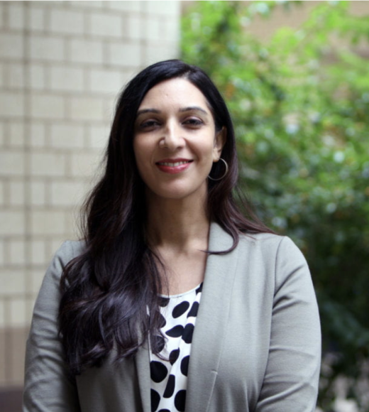 portrait of woman with long black hair, drapes over her right shoulder. She is wearing a black and white shirt, a grey sports coat and red lipstick. The corner of a tan, brick building and a green hedge are in the background.