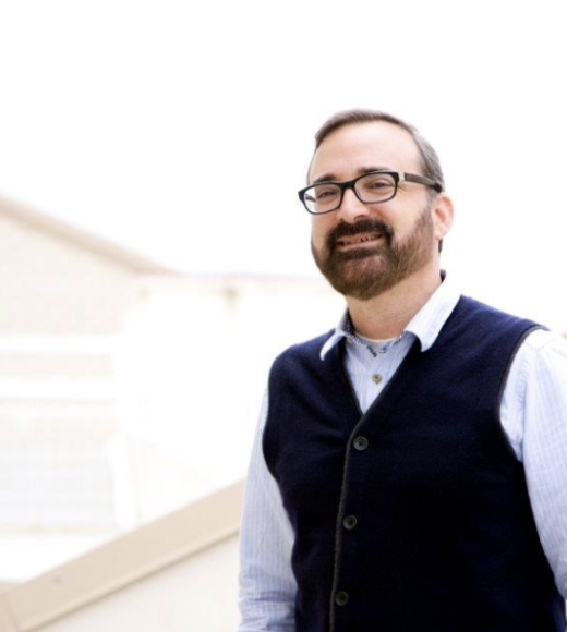 white man with dark brown hair, full beard and mustache stands in a well-lit room. He is wearing a light blue, long sleeved shirt, a navy blue sweater vest, and dark rimmed glasses.