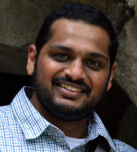 portrait of young man with dark, olive skin with short, black hair, closely cut beard and mustache smiles at camera. He is wearing a light blue checker-lined, collared shirt. 