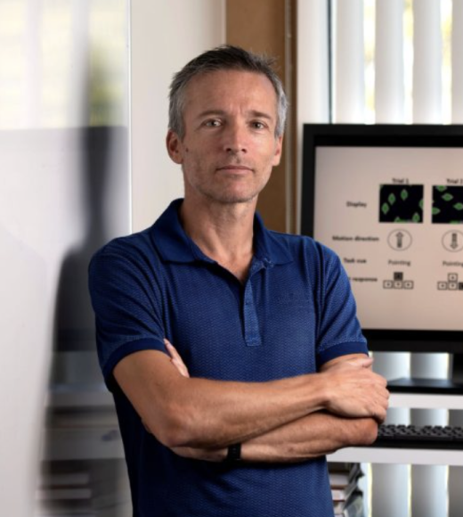 Thin white man with short grey hair stands with arms crossed, leaning against a clean whiteboard, looking at camera. He is wearing a navy blue polo and a computer is behind him. 
