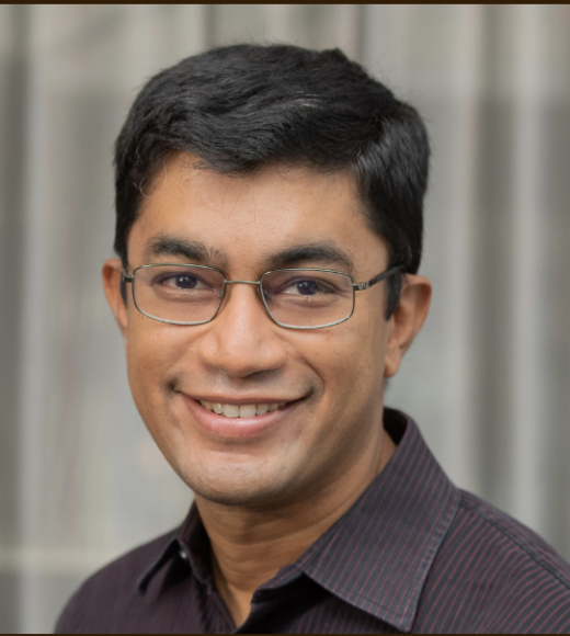 Portrait of man with black hair, olive skin smiles at camera. He is wearing thin rimmed glasses and collared shirt that is dark blue with thin, red, vertical stripes.