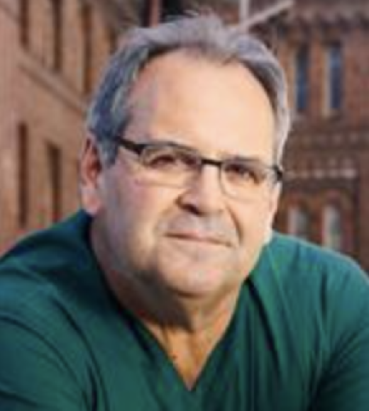 portrait of older man with grey hair looks at camera wearing horn-rimmed glasses and a green polo shirt