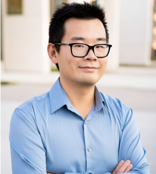 Portrait of William Wang. Young Asian man wearing dark rimmed glasses and wearing a light blue, long sleeved, collared shirt. His arms are crossed.