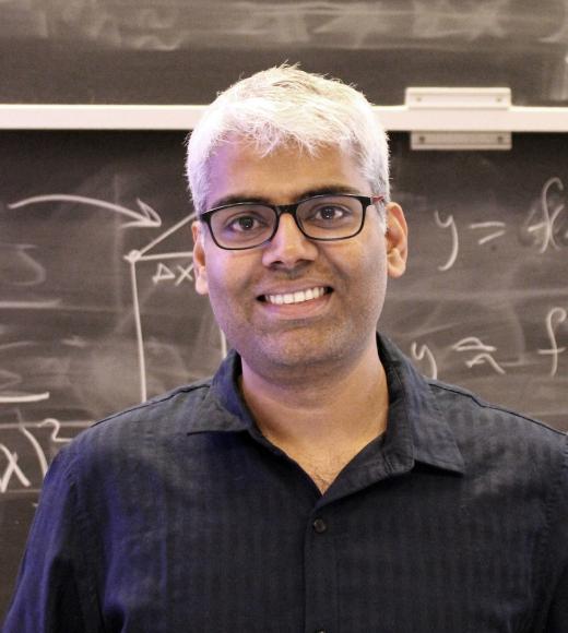 Gopala Krishna Anumanchipalli stands in front of a chalk blackboard with math equations on it. He has short, white hair, glasses, olive skin and is wearing a black collared shirt 