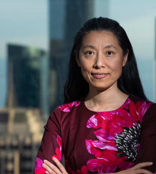 Asian woman with long black hair wearing a dress with large, brightly colored pink flowers printed on it, stands with arms crossed front of a cityscape.