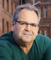 portrait of older man with grey hair looks at camera wearing horn-rimmed glasses and a green polo shirt