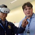 Man wearing virtual reality head set points finger in the air with a male graduate student standing nearby looking at a laptop to guide the interaction.