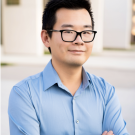 Portrait of William Wang. Young Asian man wearing dark rimmed glasses and wearing a light blue, long sleeved, collared shirt. His arms are crossed.