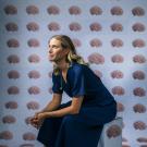 Emily Jacobs sits on a box with hands clasped resting on her knees. She is looking up off to the side of the image. Behind her and covering the box is wall paper that has rows and columns of human brains on a light blue background.