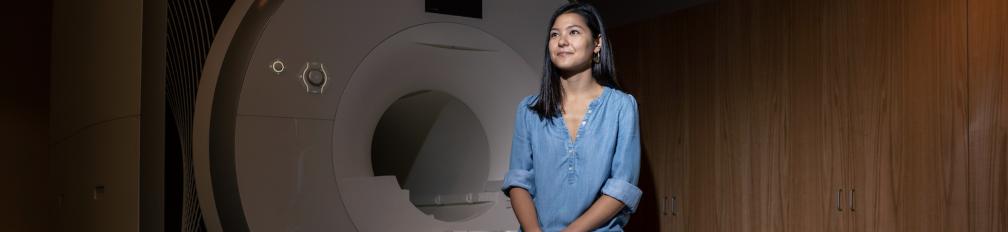 Olive-skinned woman with long black hair, wearing a light blue shirt sits in front of an MRI scanner. She is smiling looking off into the distance. 